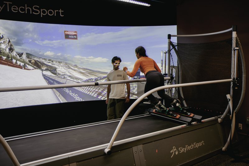 Two people using a SkyTechSport Ski Simulator with large curved mountain display screen in an indoor facility.