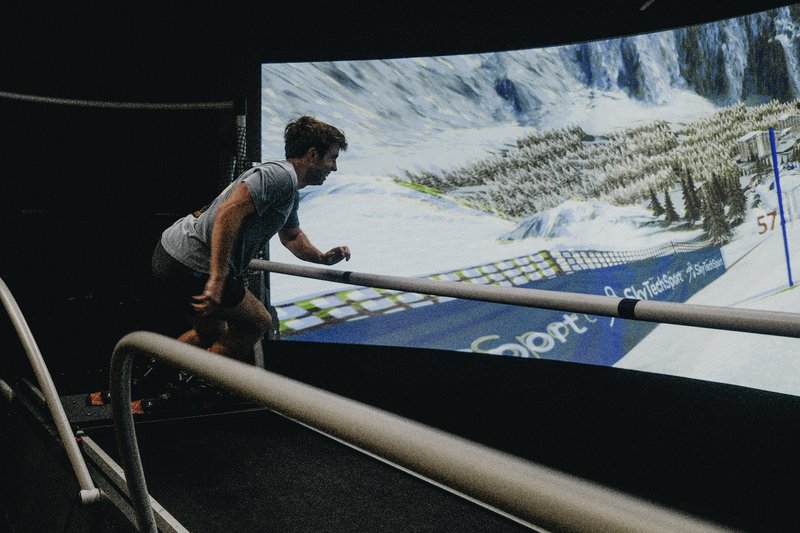 A person wearing VR goggles uses the SkyTechSport Ski Simulator with immersive alpine slope visuals displayed on a large curved screen.
