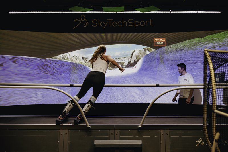 A woman uses the SkyTechSport Ski Simulator while an instructor observes, with snowy mountain scenery displayed on the large curved screen behind them.