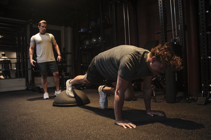 A person performs a plank exercise with feet elevated on a BalancePlay Pro Pro while a trainer observes in a modern fitness facility.
