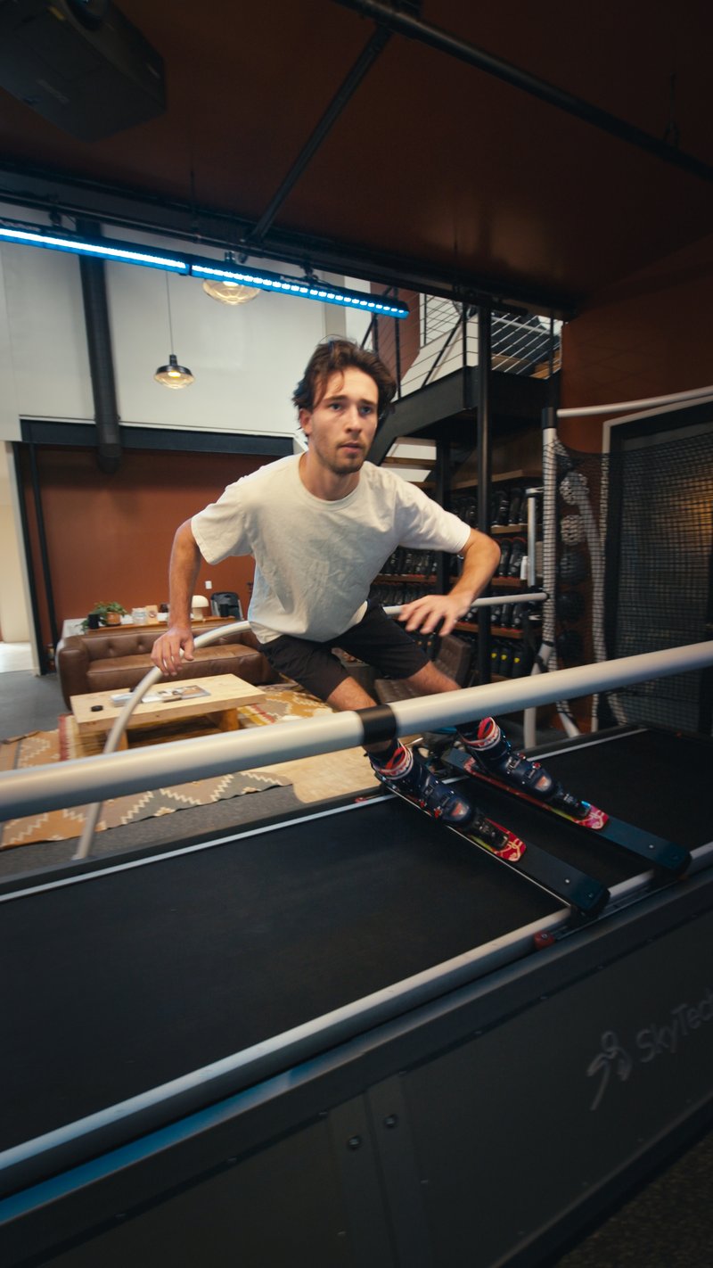 A young man in a white t-shirt sits beside a SkyTechSport Ski Simulator in a modern indoor facility with industrial design elements.