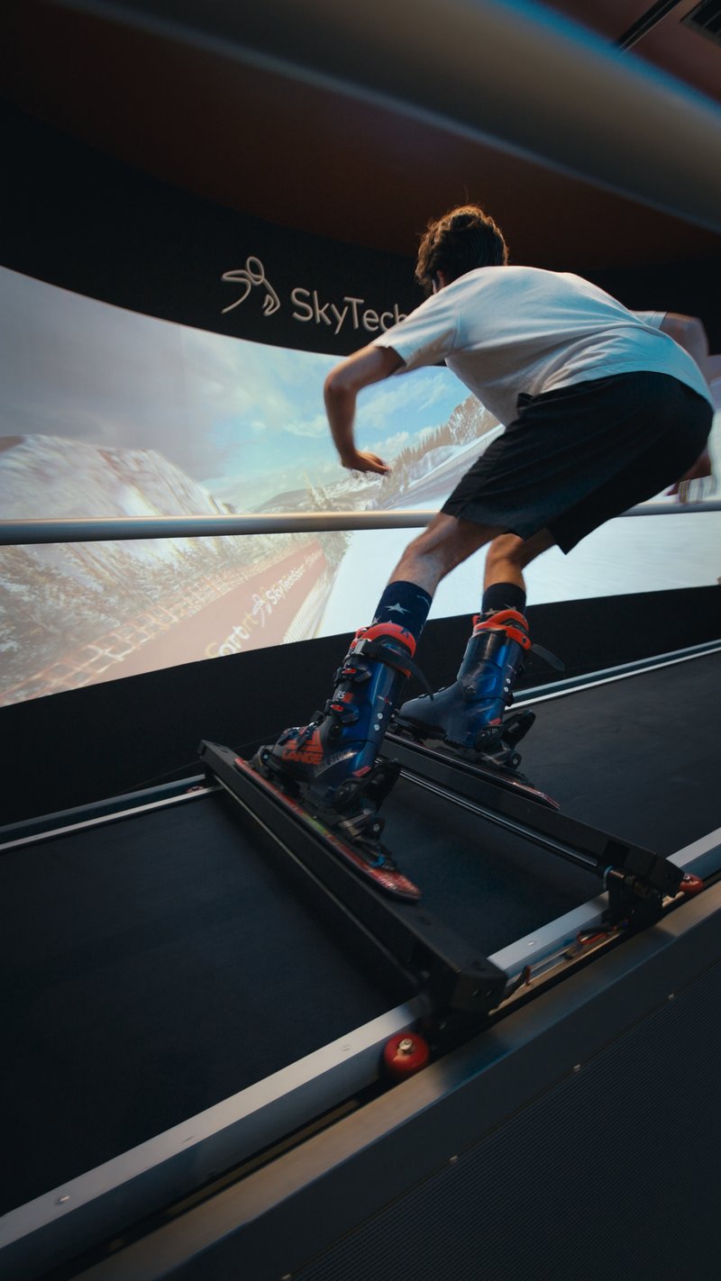 A person in ski boots practices skiing technique on a SkyTechSport Ski Simulator with immersive mountain scenery displayed on the screen.