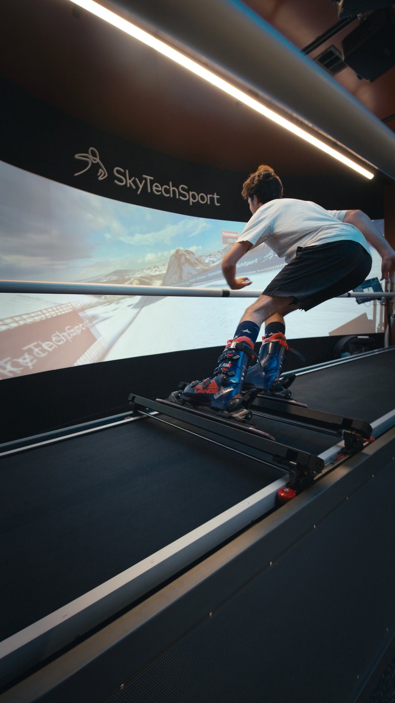 A skier in racing position practices on the SkyTechSport Ski Simulator with an immersive mountain landscape display in the background.