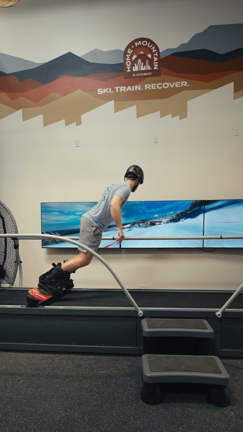 A person using the SkyTechSport Ski Simulator in a modern fitness facility with mountain-themed wall graphics.