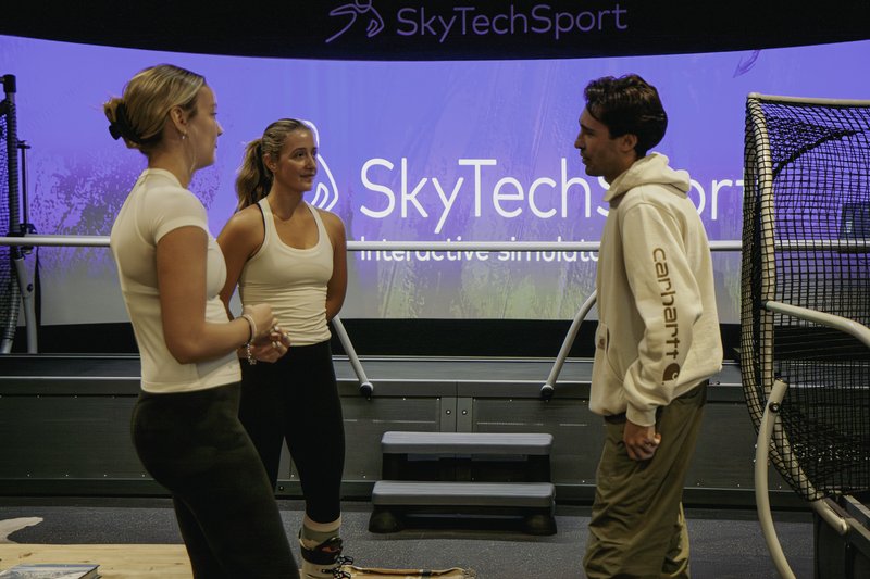 Three young adults — two women in athletic wear and a man in a Carhartt hoodie — chat casually in front of a large curved SkyTechSport interactive simulator screen inside the Home Mountain facility.