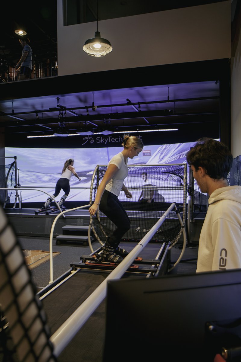 Two athletes train on SkyTechSport Ski Simulators inside the Home Mountain facility, with an immersive projected ski slope screen displaying the SkyTechSport logo behind them while a third person observes from the mezzanine level above.
