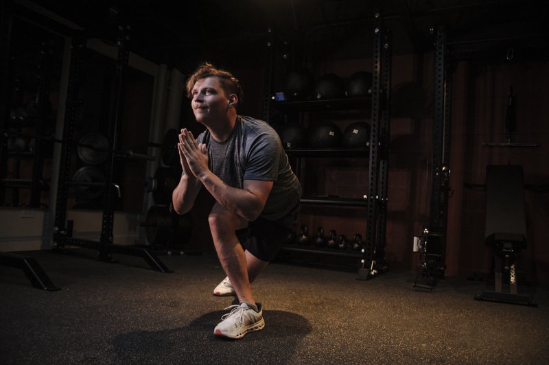 A young man performs a deep squat exercise in a well-equipped gym with weights and equipment in the background.