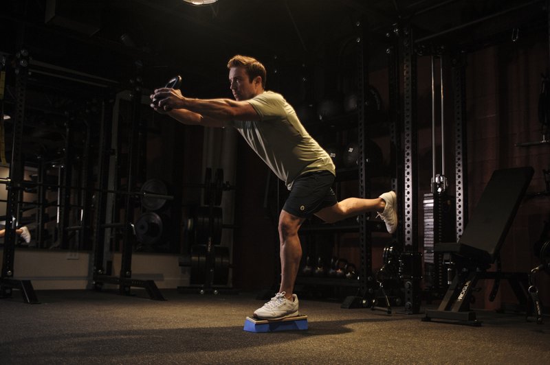 A man in athletic wear performs a dynamic lunging exercise while holding weights on a blue balance platform in a modern gym setting.