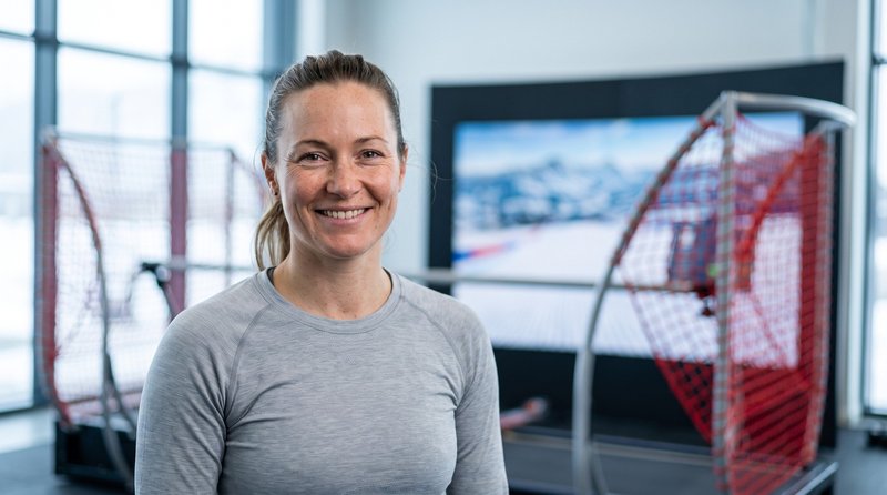 A smiling woman in a grey athletic top poses confidently inside the Home Mountain facility, with a SkyTechSport Ski Simulator and a large ski-slope screen visible in the background.