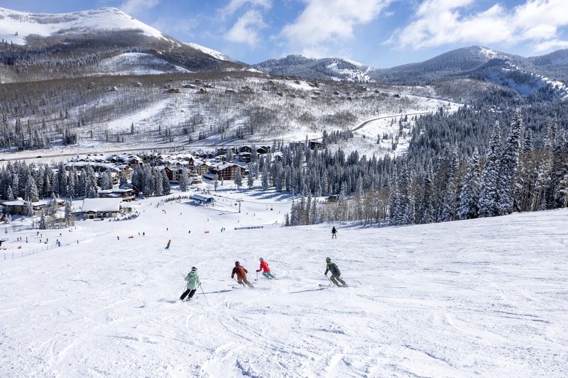 Four skiers descend a groomed run together at a mountain resort, with snow-covered pines, a village base area, and dramatic peaks filling the background under bright winter skies.