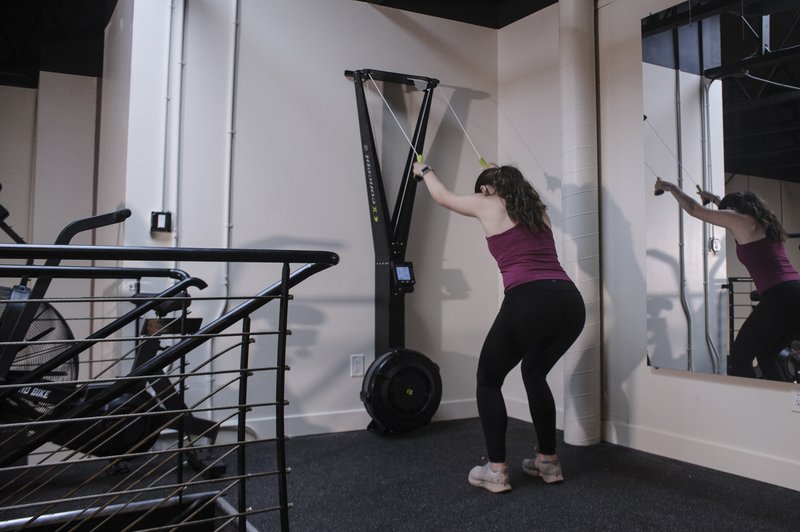 A woman in purple top and black leggings performs resistance training exercises with a wall-mounted pulley system in a modern indoor gym facility.