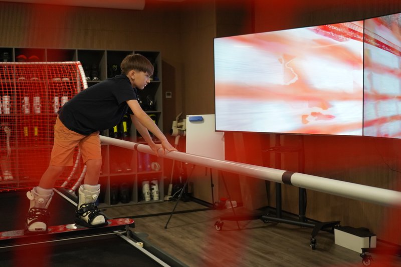 A young person uses the SkyTechSport Ski Simulator with ski boots and poles while a large screen displays snowy mountain scenery.