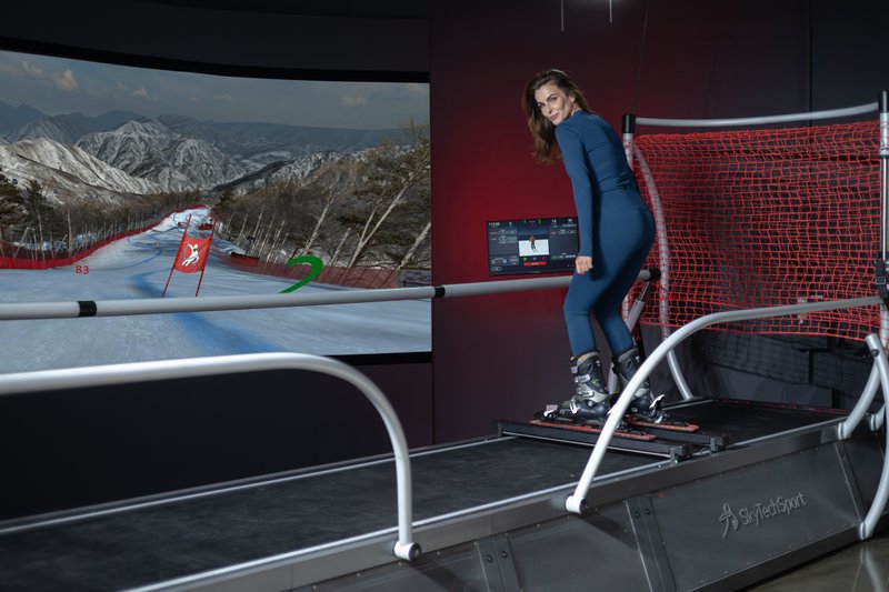 A woman in blue ski attire demonstrates the SkyTechSport Ski Simulator with a large curved mountain skiing display screen in the background.