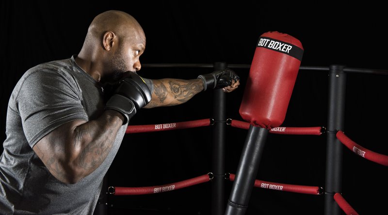 A muscular athlete wearing MMA gloves throws a punch at a red BotBoxer robotic training target mounted on a black frame with red ropes, set against a dark studio background.