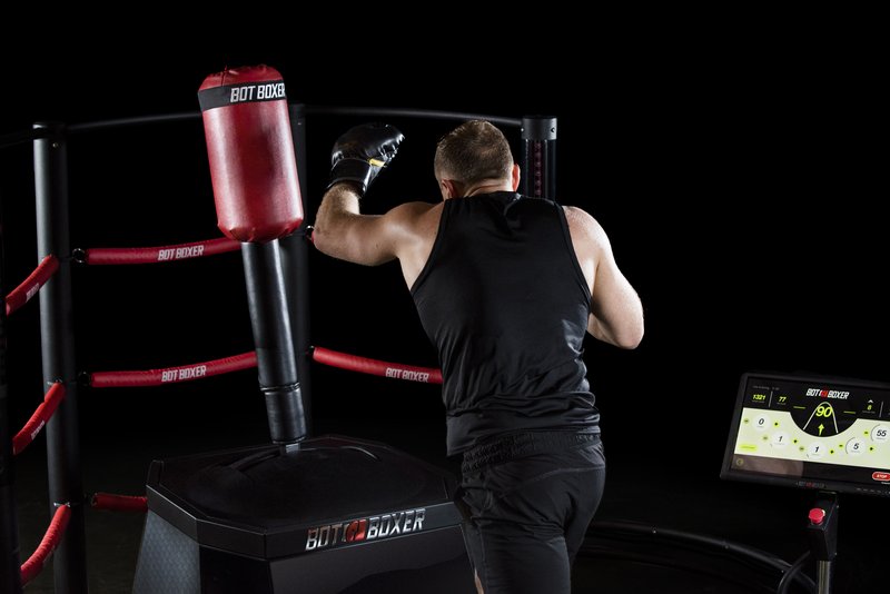 A male athlete in black workout attire throws a punch at a BotBoxer robotic BotBoxer, with the branded frame, red ropes, and performance display screen visible against a dark studio background.