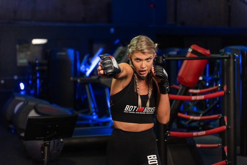 A female athlete wearing BotBoxer-branded sportswear and MMA gloves throws a punch in a darkened gym setting with BotBoxer training equipment visible in the background.