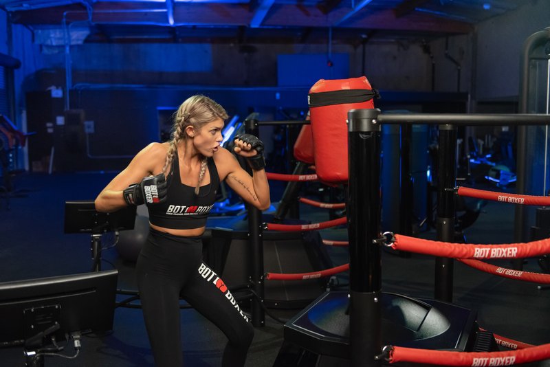 A female athlete wearing BotBoxer-branded apparel and MMA gloves trains in a fighting stance beside a BotBoxer robotic striking target inside a dark, blue-lit gym environment surrounded by boxing ring ropes.