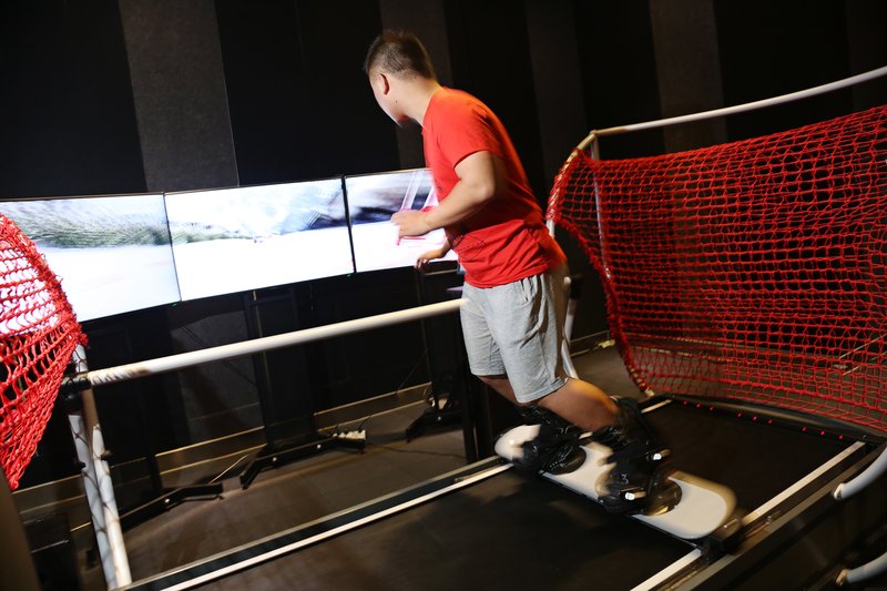 A man in a red shirt and grey shorts stands on the moving platform of a SkyTechSport Ski Simulator, gripping the red safety rail netting while facing a multi-panel immersive ski-slope screen in a darkened indoor installation.
