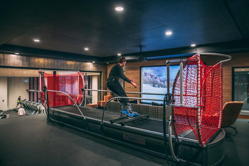 A man in ski boots carves dynamic turns on a SkyTechSport Ski Simulator inside an upscale indoor training facility, with red safety netting on both sides, a front-mounted projection screen displaying an alpine snow scene, and a mirrored gym visible in the background.