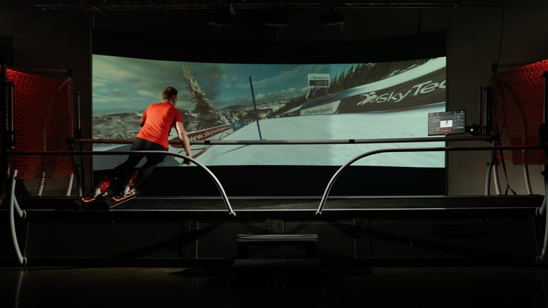 A skier in a red top trains on the SkyTechSport Ski Simulator in a darkened indoor installation, facing a large curved immersive projection screen displaying a alpine ski race course with SkyTech branding visible.