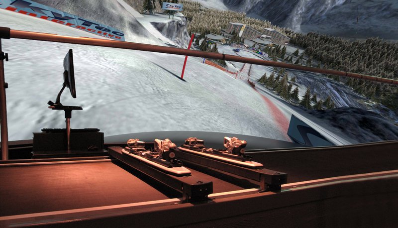 A close-up view from inside the SkyTechSport Ski Simulator cockpit showing ski bindings mounted on the motion platform rail in the foreground, with a giant panoramic projection screen displaying an immersive alpine slalom course with snow-covered slopes, trees, and ski resort infrastructure.