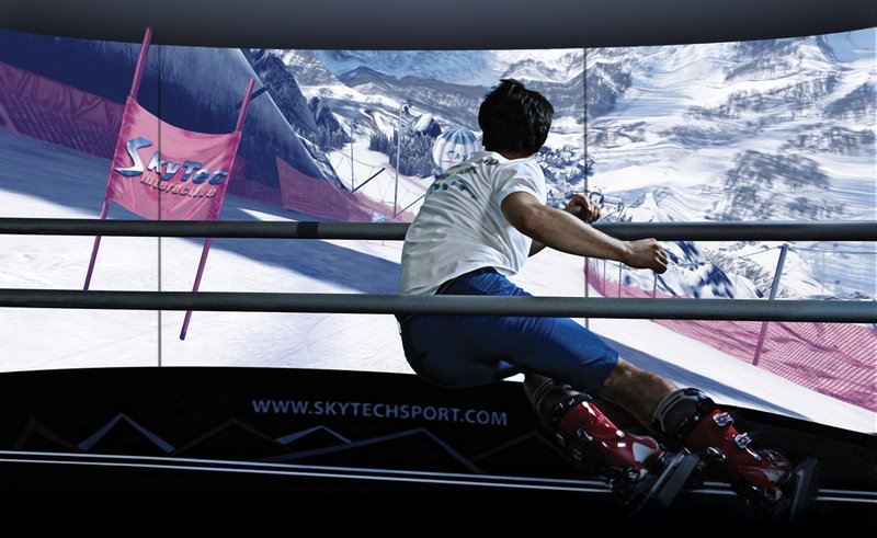 A male skier in ski boots and racing posture rides the SkyTechSport Ski Simulator on a dynamic motion platform, fully immersed in a curved panoramic giant-slalom race course projection bearing the SkyTechSport logo and website URL.